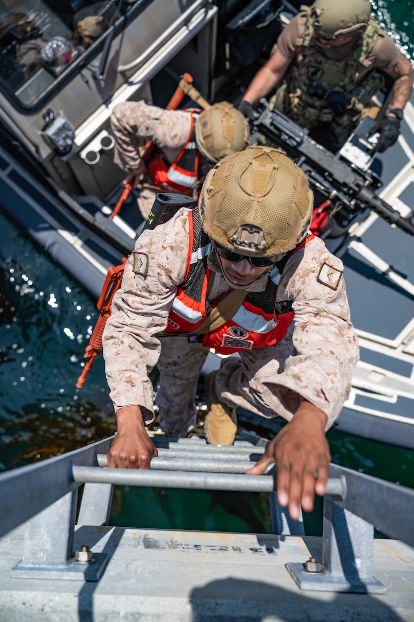 Marines climb up a ladder from a small boat wearing helmets on a sunny day as seen from above.