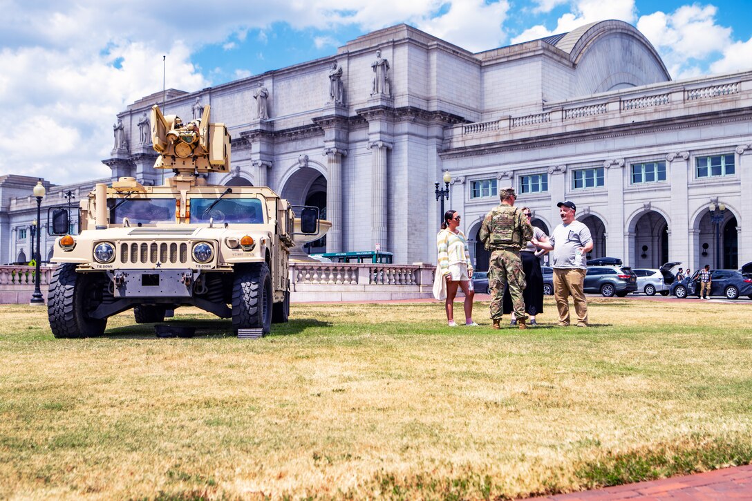 A soldier talks to members of the public while standing near a Humvee parked on grass in front of a large building, with other vehicles and people in the background on a cloudy day.