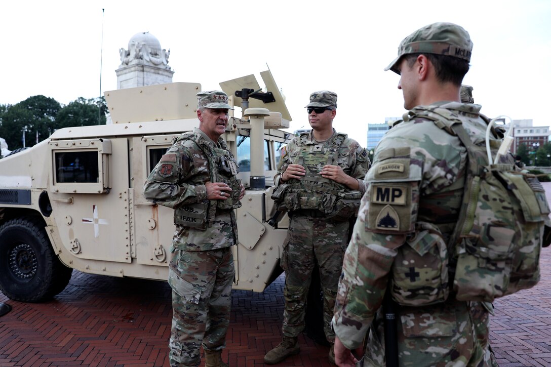 An airman stands and talks to three soldiers on a brick patio, with a Humvee parked behind them and a monument and trees in the background on an overcast day.