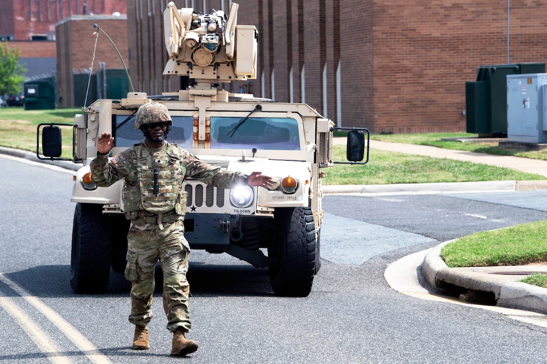 A guardsman walks along a paved road and uses hand signals to guide a Humvee that is being driven behind the soldier, with buildings, grass and a sidewalk in the background on a sunny day.