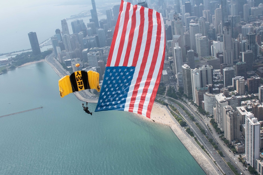 A soldier descends with a parachute that says "Army" while displaying a large American flag over a city's coastline.
