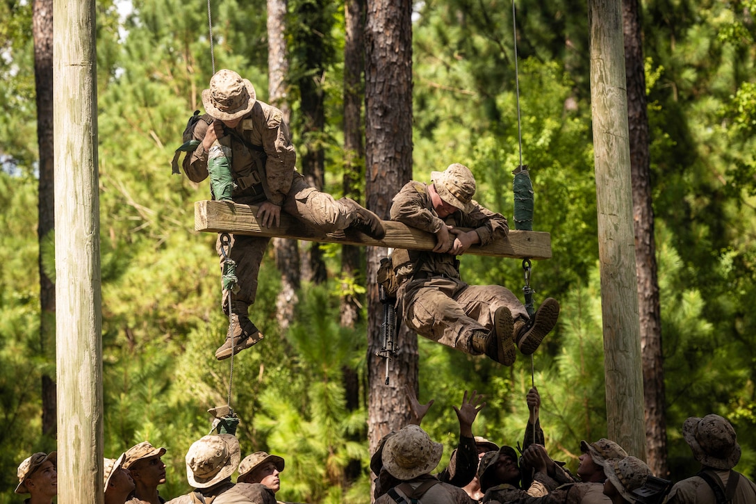Two Marine Corps recruits climb a wooden structure as dozens of fellow recruits provide support from below in a wooded area.