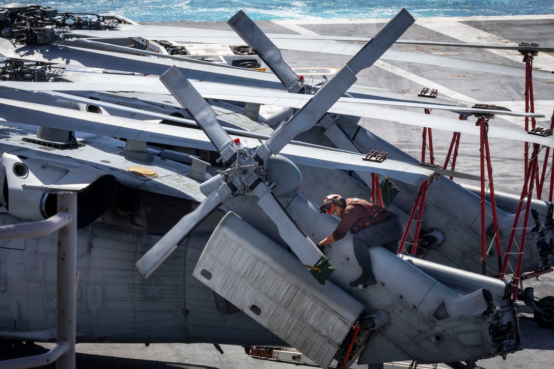 A sailor holds onto a helicopter while performing maintenance on it aboard a ship at sea.