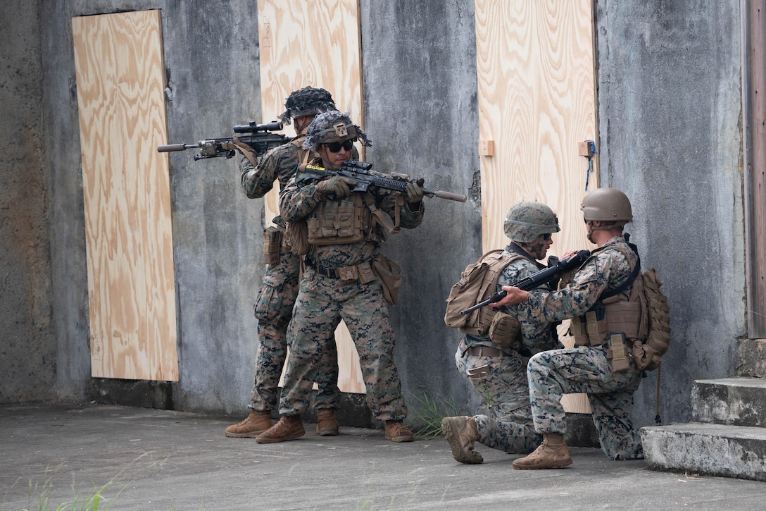 Two Marines kneel next to a wooden door while two others stand watch with weapons in a concrete structure.