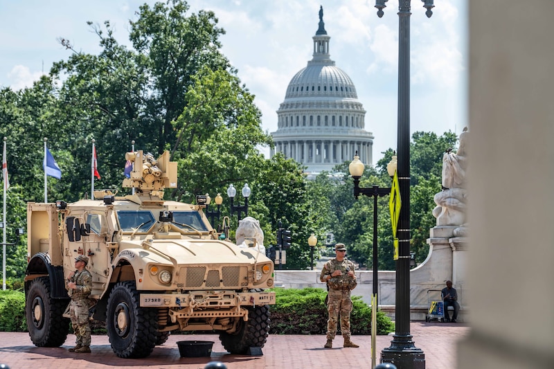 Two soldiers stand next to a military vehicle with a monument in the background on a sunny day.