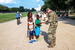 An airman hands something to a child as another child and parent watch on a rocky surface near a monument on a sunny day.