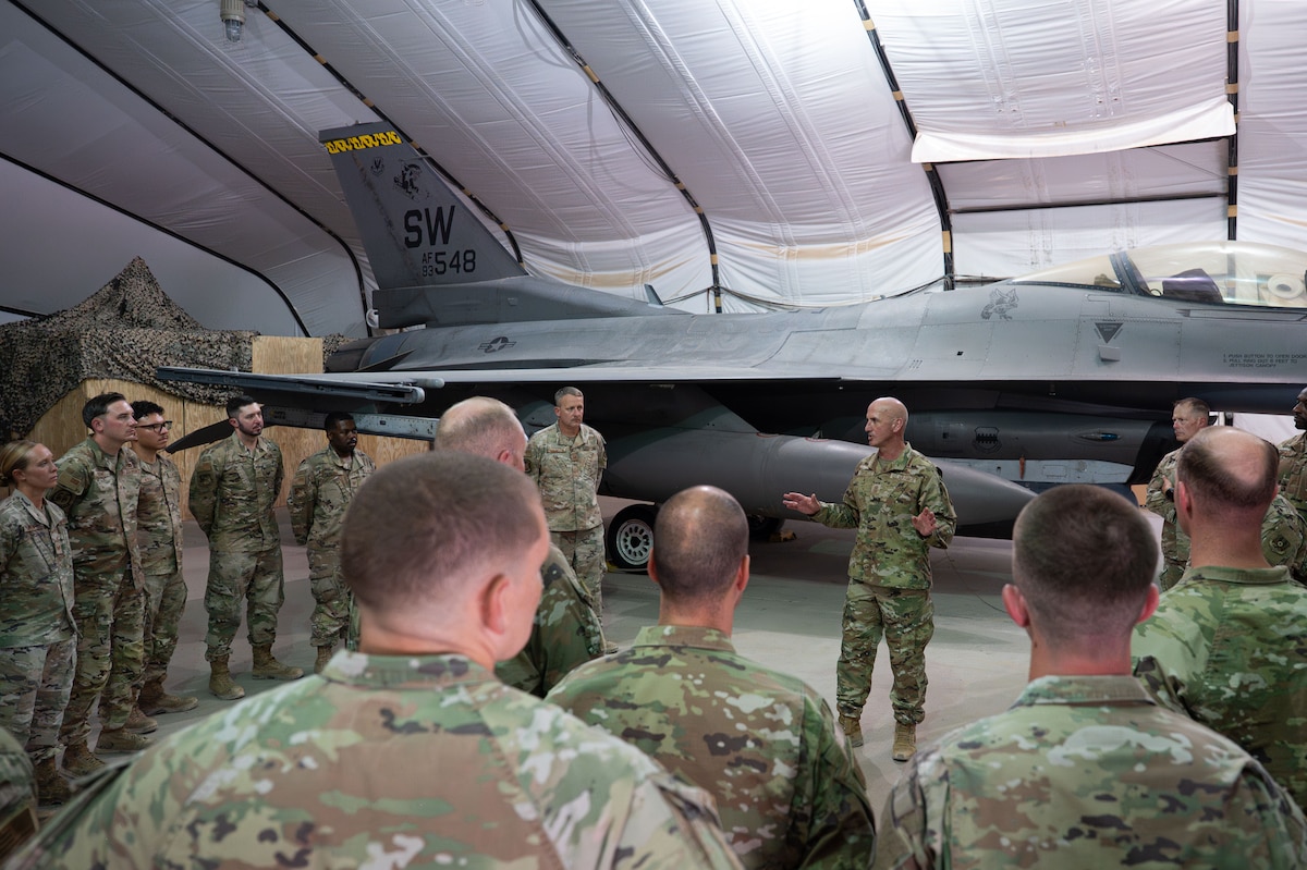 A man talks to a group of people in front of an F-16 Fighting Falcon.