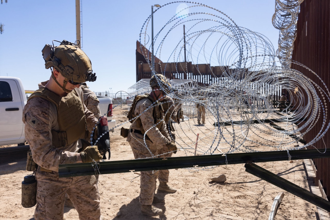 Two people wearing camouflage military uniforms place concertina wire onto brackets of barrier fence in a desert environment.