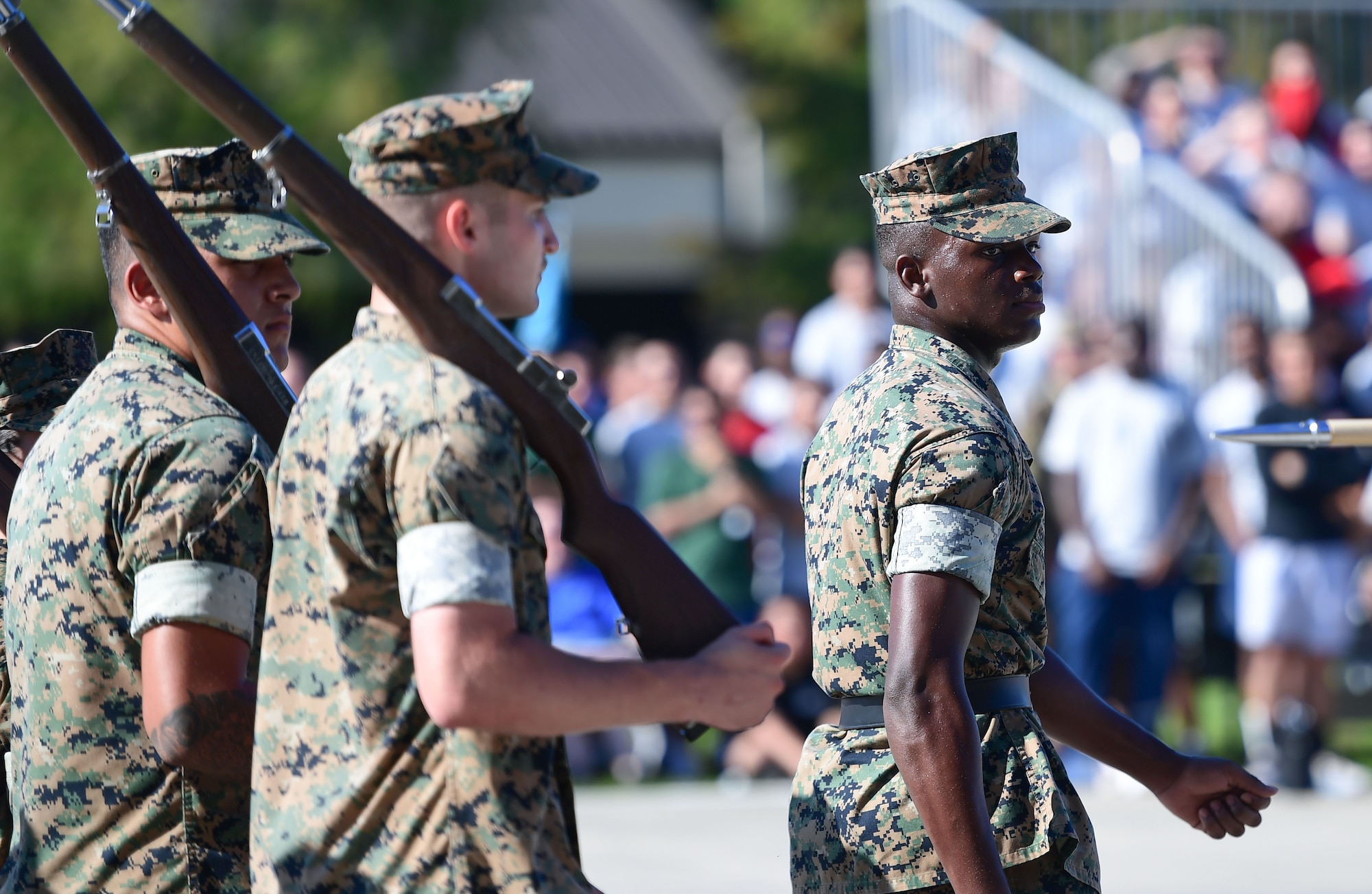 Men in military uniform march in formation while holding fake rifles.