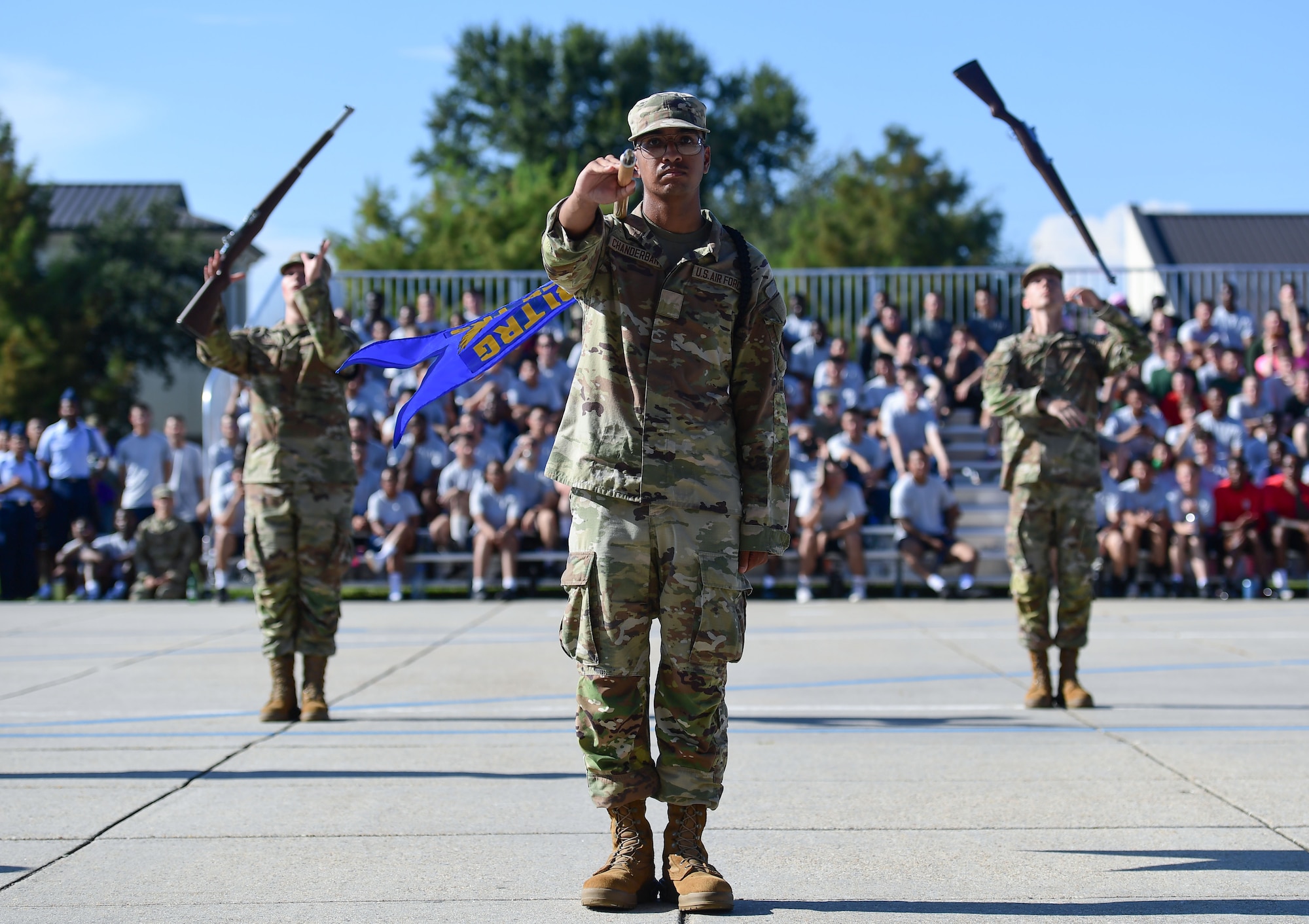 A man in military uniform stands center while holding a flag on a stick, while two men in the background spin static weapons in the air.