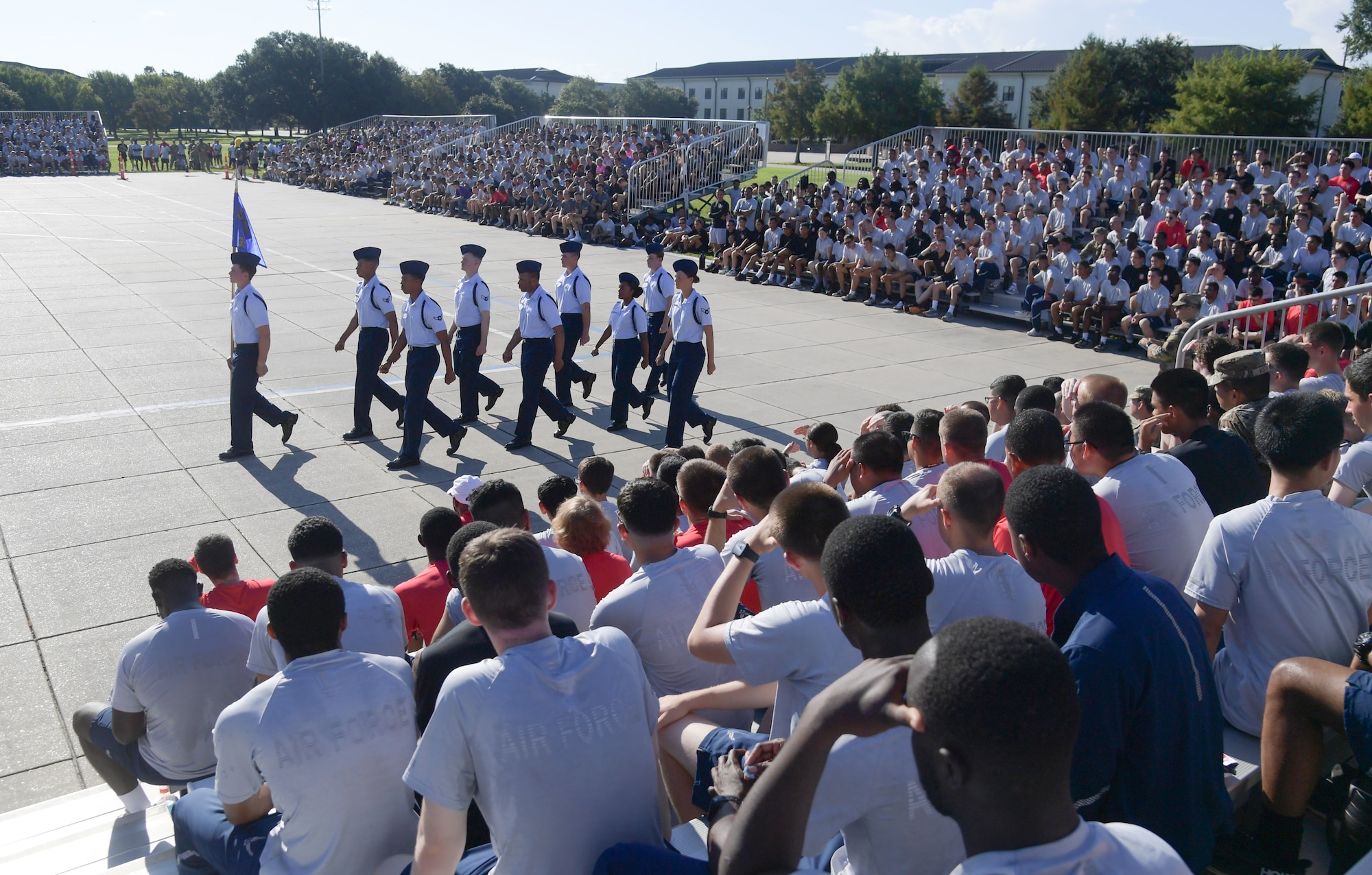 A group of people wearing military uniform march in formation in front of bleachers full of people.
