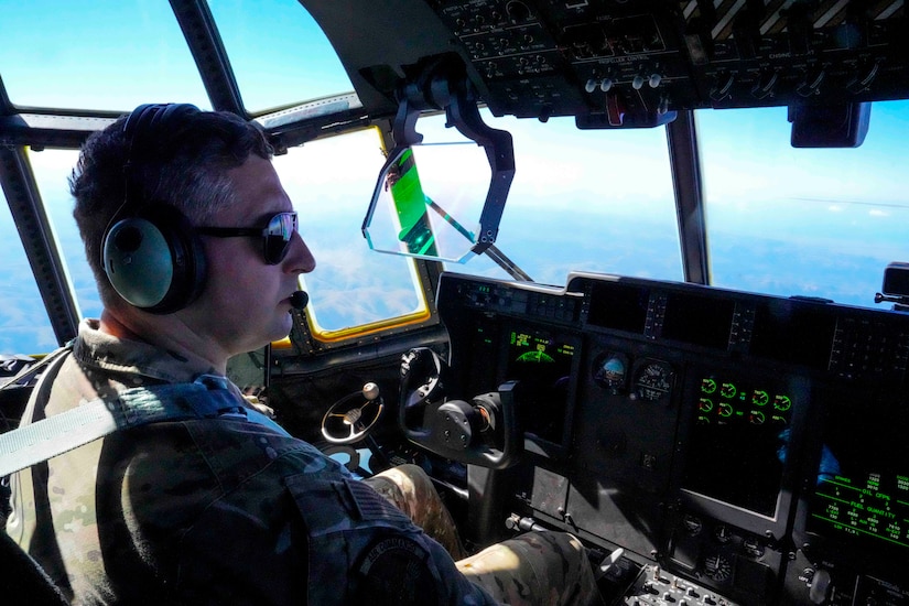 An airman pilots an aircraft as seen from the cockpit with a blue sky and a view of the ground is shown through the windows.