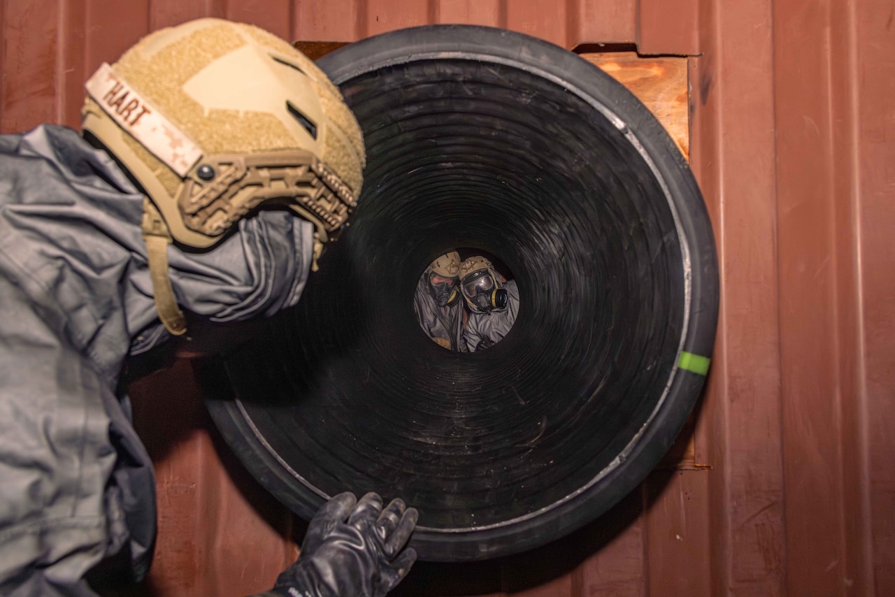 A Marine in a helmet looks down a large tube at two Marines on the other side.