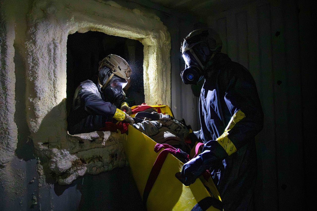 Two Marines in hazmat suits and masks carry a simulated casualty through an opening in a dark room.