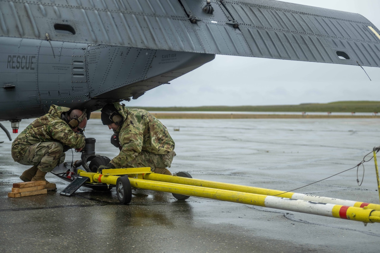 Two guardsmen crouch over yellow equipment under an aircraft outside on a runway on a cloudy day.