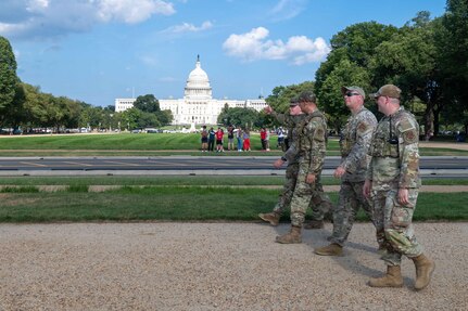 Four people wearing camouflage military uniforms walk down the street in front of the U.S. Capitol. There are people in the background, along with trees and a blue sky with white clouds.