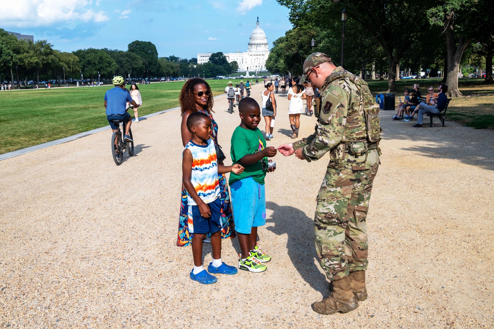 An airman hands something to a child as another child and parent watch on a rocky surface near a monument on a sunny day.