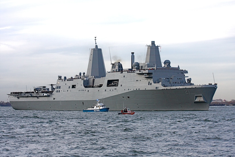 A large gray military ship sails on a river beside a tugboat and a patrol vessel.