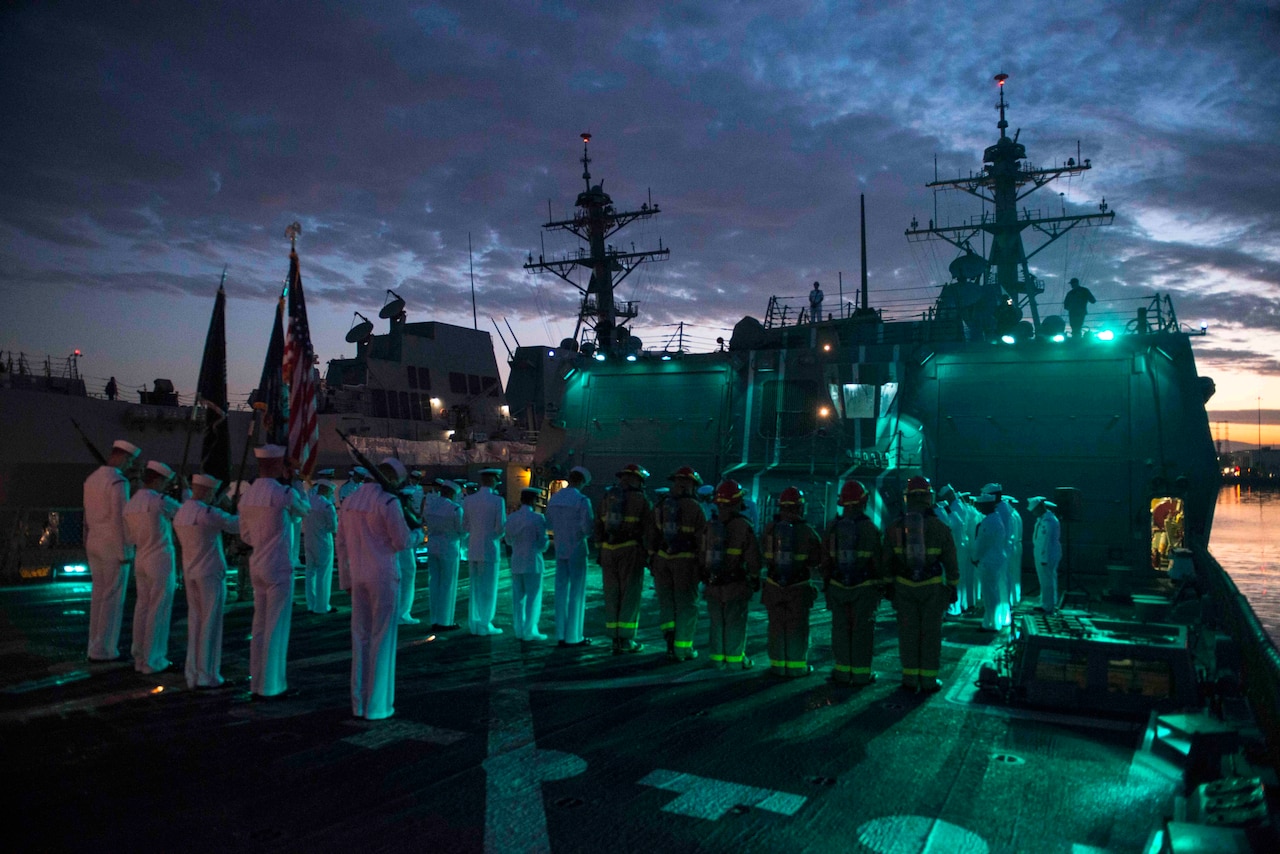 Sailors in military dress uniforms and firemen aboard a ship at night bow their heads. The sky is cloudy and there is another large gray ship in the background.