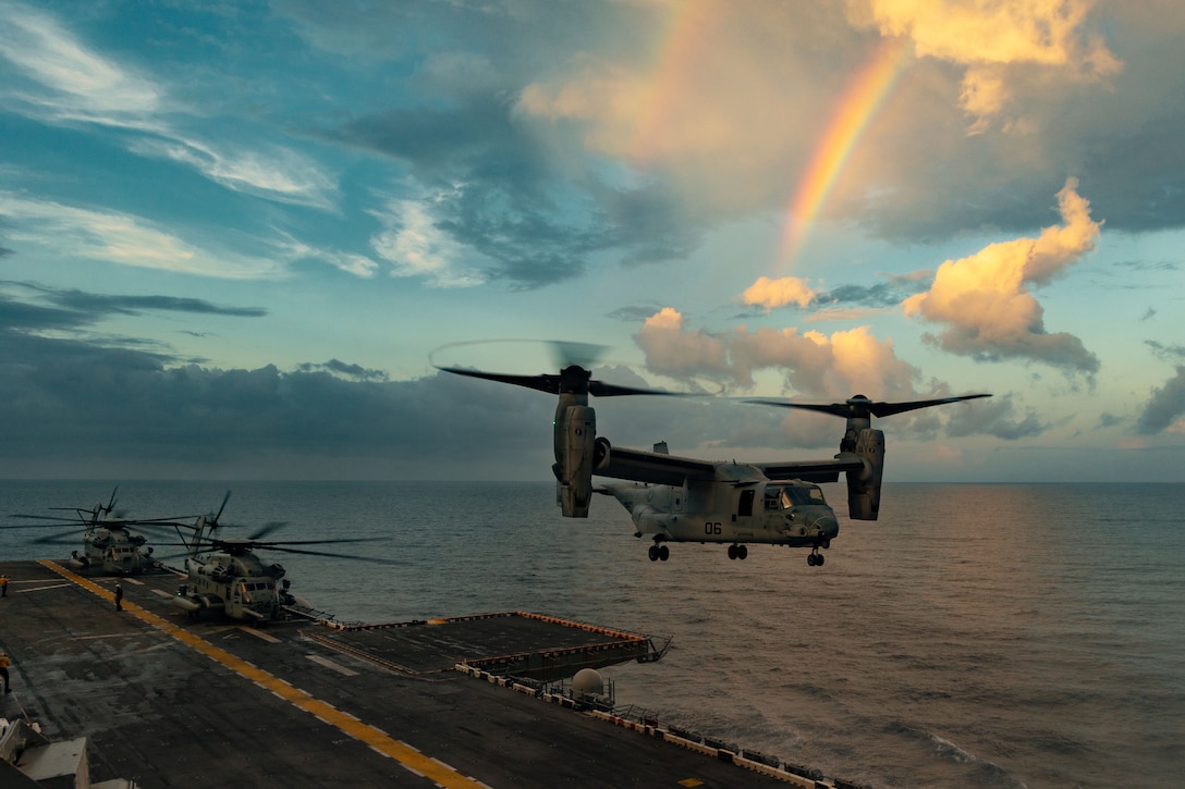 A Marine Corps Osprey hovers above a ship at sea, with a rainbow in a cloud-streaked sky in the background.
