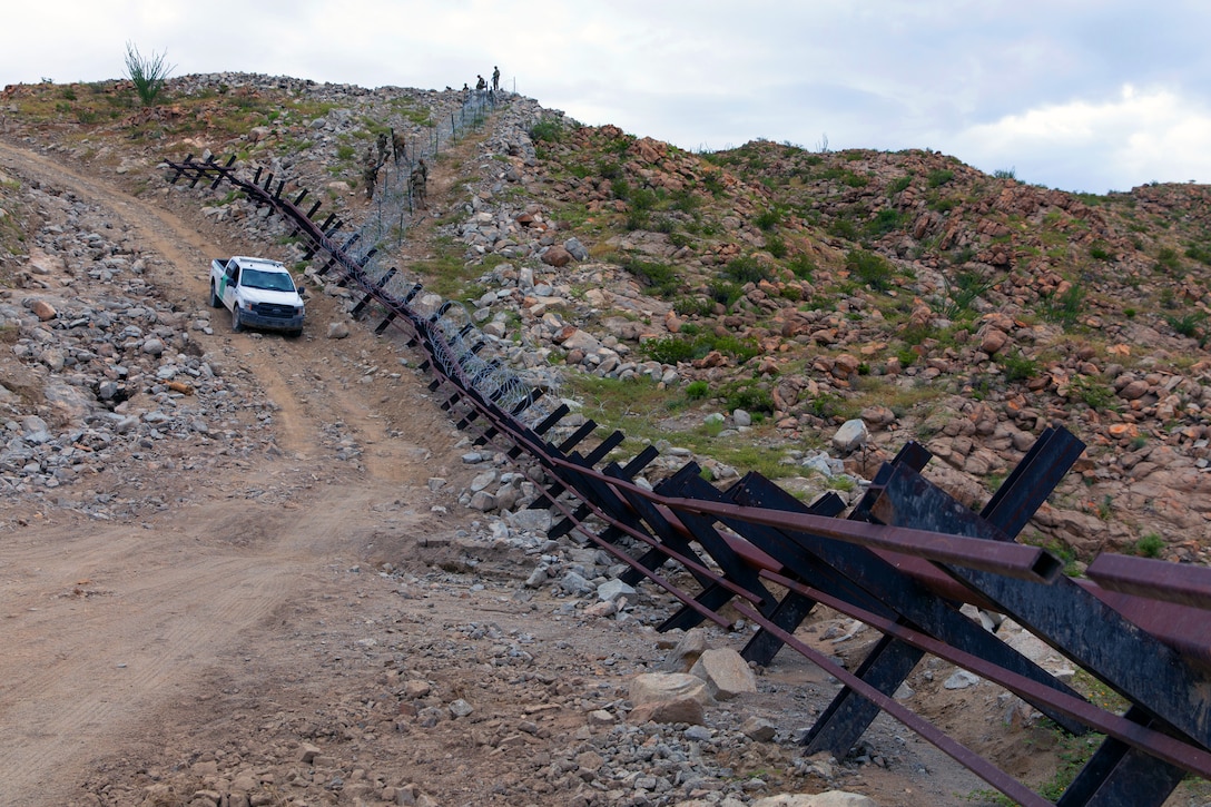 Soldiers in the distance walk down a hill towards an obstacle next to a road as a truck drives by.