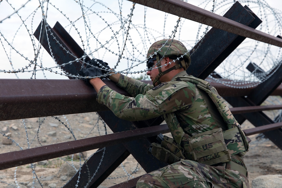 A soldier leans over and secures concertina wire to an obstacle on a cloudy day.