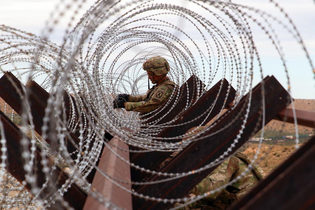 A soldier installs concertina wire on a wooden fence outside as seen through the wire.