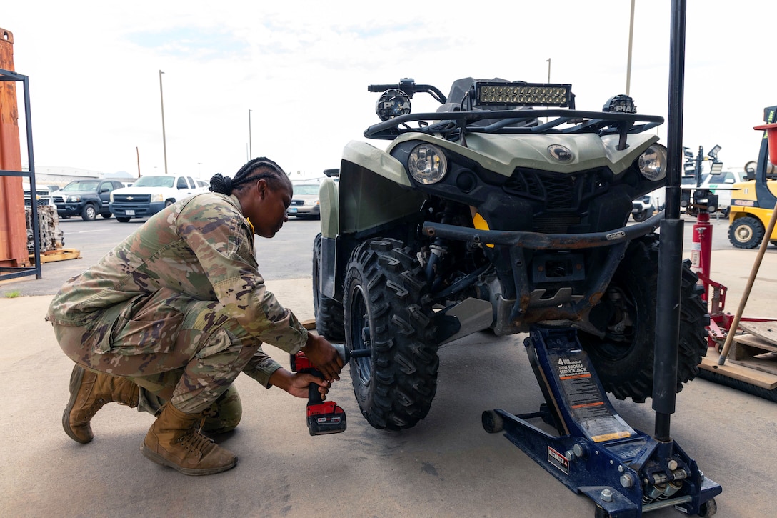 A solider crouches next to an ATV to install a tire using a red tool outside in a parking lot.