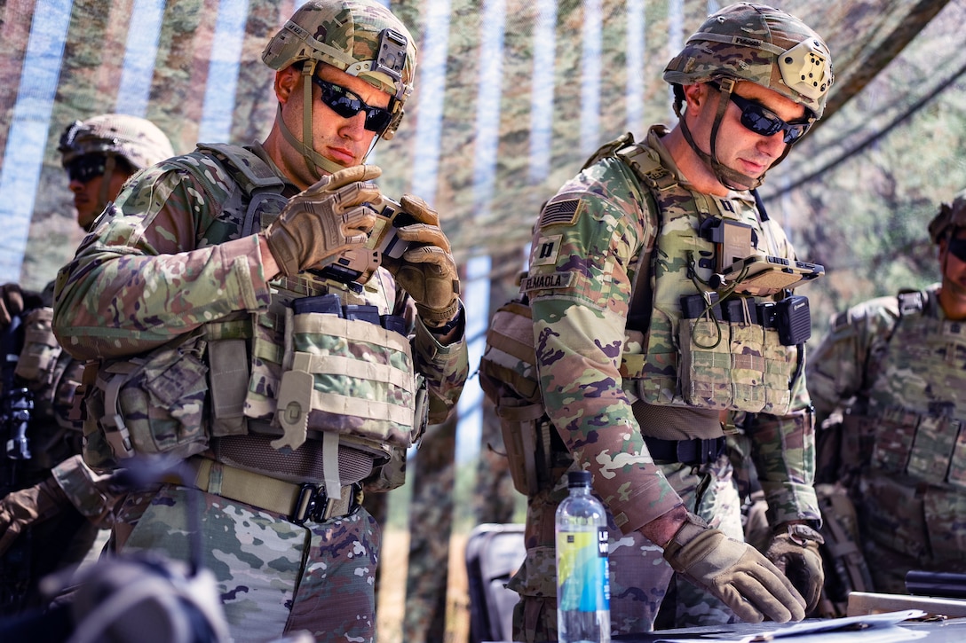 Two soldiers in tactical gear and sunglasses stand under a mesh tent near a border wall and look down at a table while other soldiers are behind.