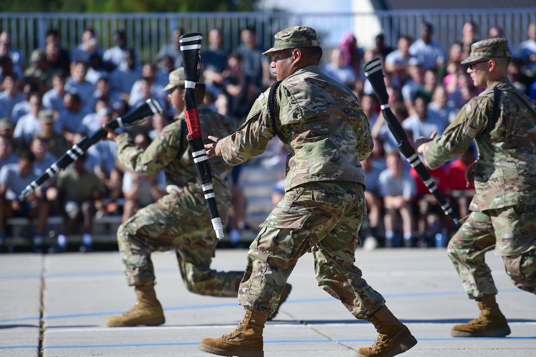 Three airmen with knees bent twirl rifles on pavement outdoors as a seated crowd watches.