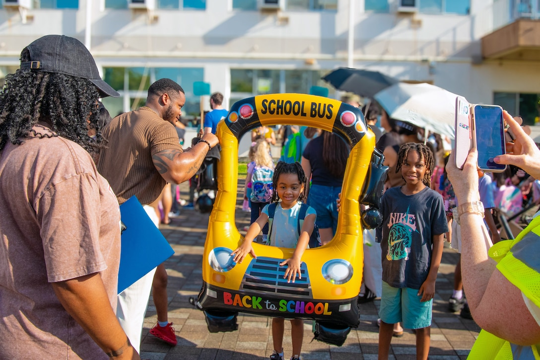 A child carrying an inflatable school bus stands with another kid as adults surround them applauding and taking photos outside a building.
