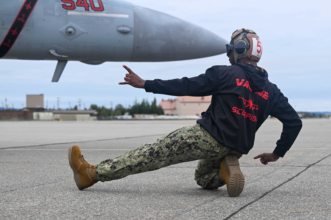 A man dressed in Navy camouflage uniform pants, a casual jacket and wearing hearing protection, guides a jet for takeoff.