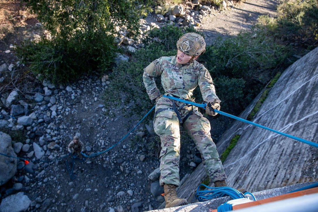 A soldier uses a blue rope to rappel down a vertical outdoor surface as another watches from rocky terrain below.