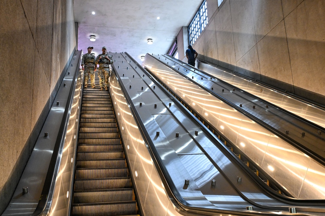 Two airmen in uniform ride a long down escalator as a civilian goes up on an adjacent escalator.