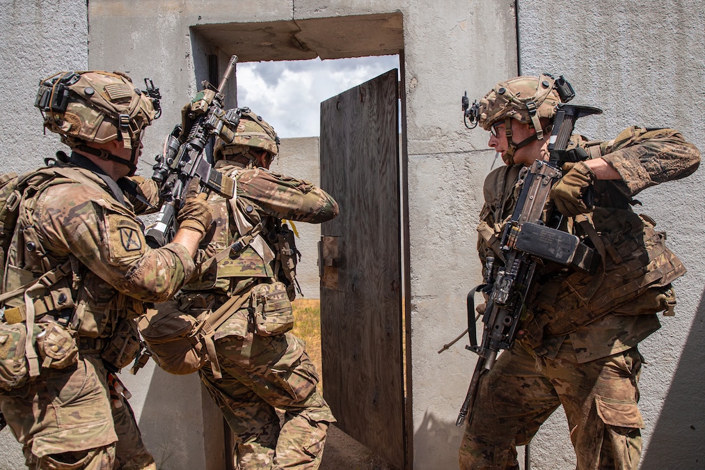 Three soldiers wield rifles while moving toward an open door on an outdoor structures.