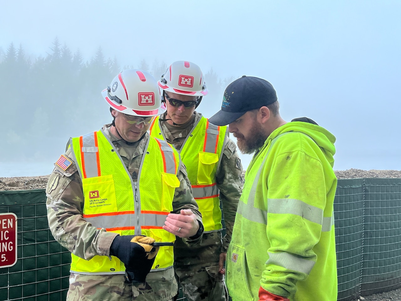 Three people, two wearing camouflage uniforms, reflective vests and helmets, and another in a safety jacket and a baseball cap, look at the screen of a smartphone.
