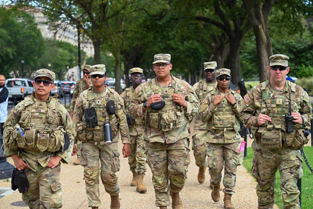Eight soldiers patrol along a paved walkway, with citizens, vehicles on a street and trees in the background.