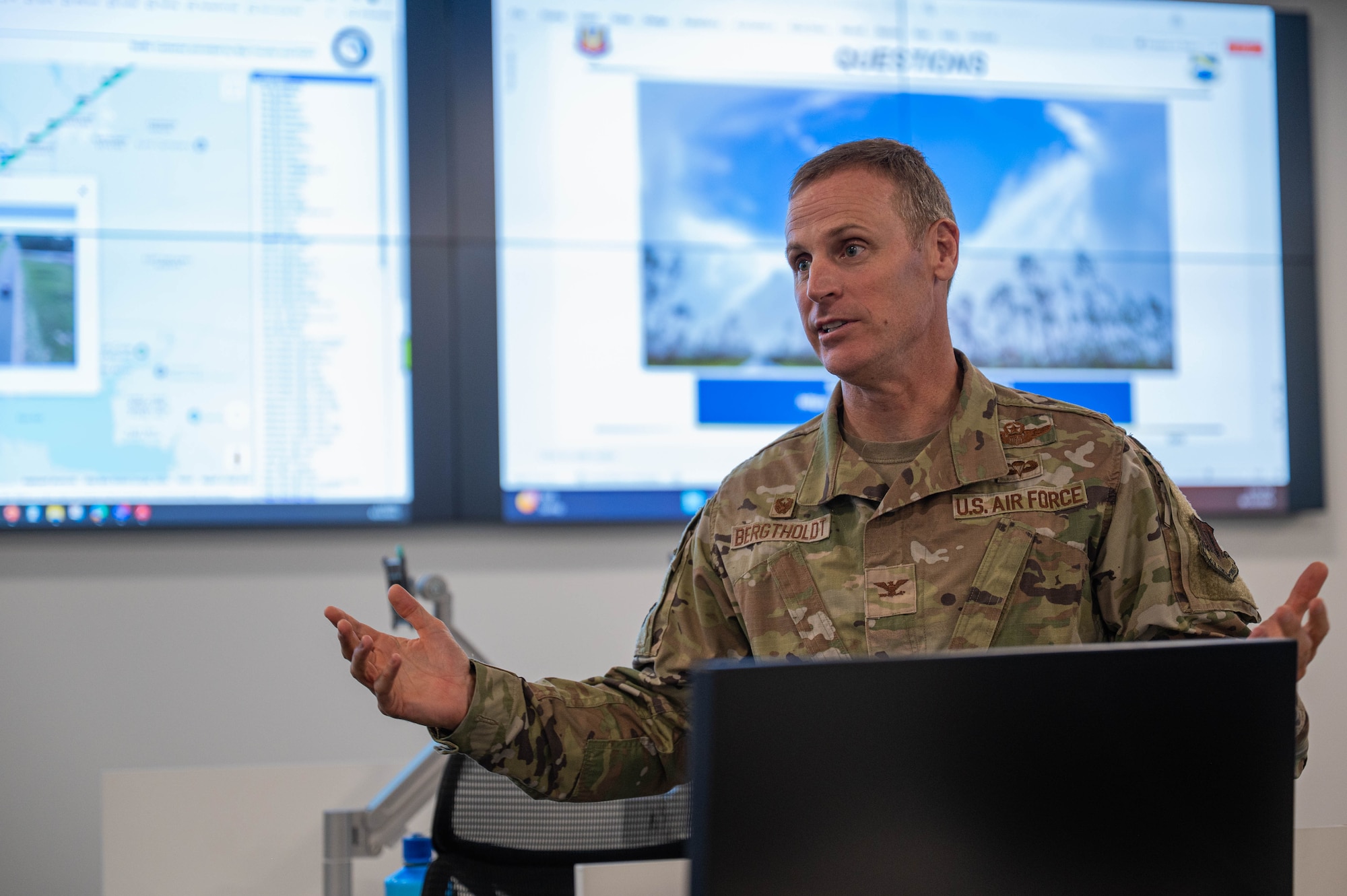 The base commander stands at the front of a room and addresses them.