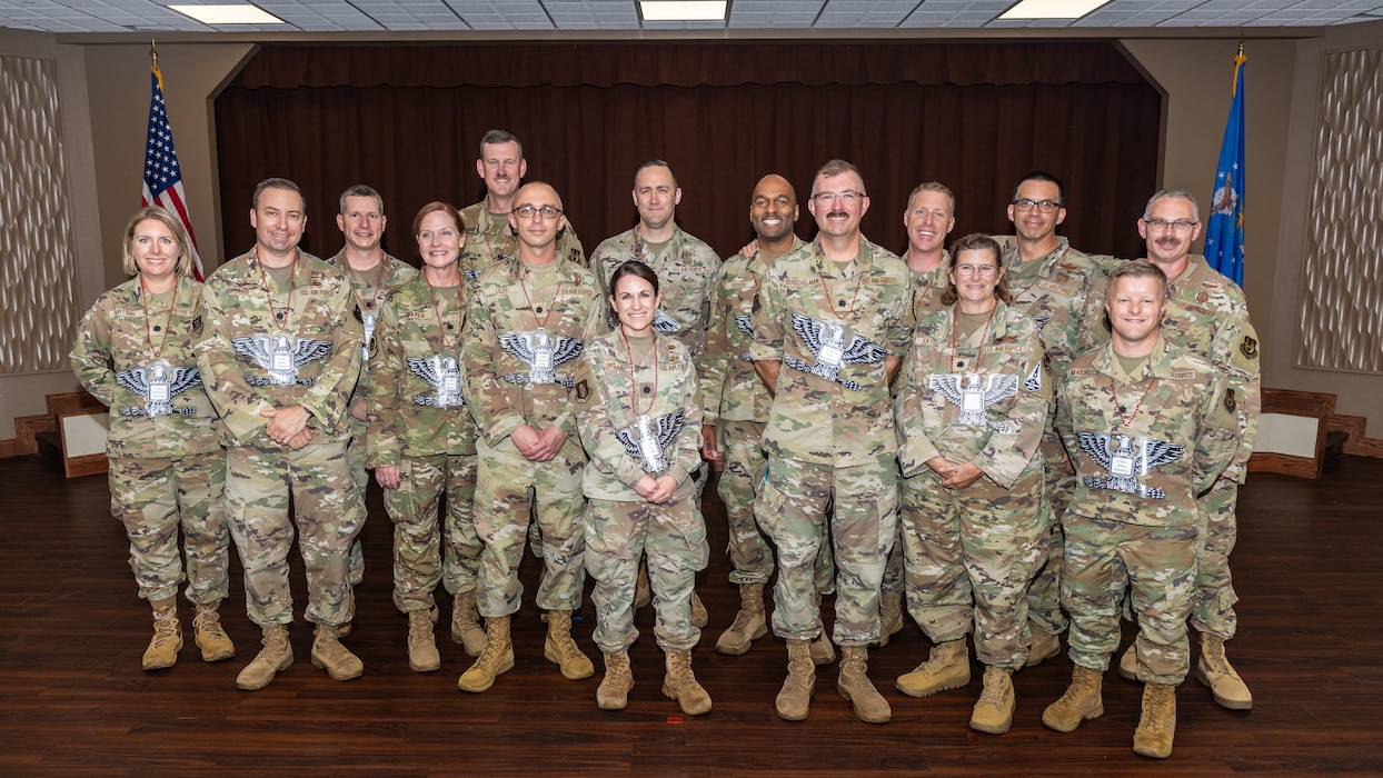 A group photo of men and women, wearing military camouflage uniforms, pose for a group photo with large, laminated colonel rank hung from their necks