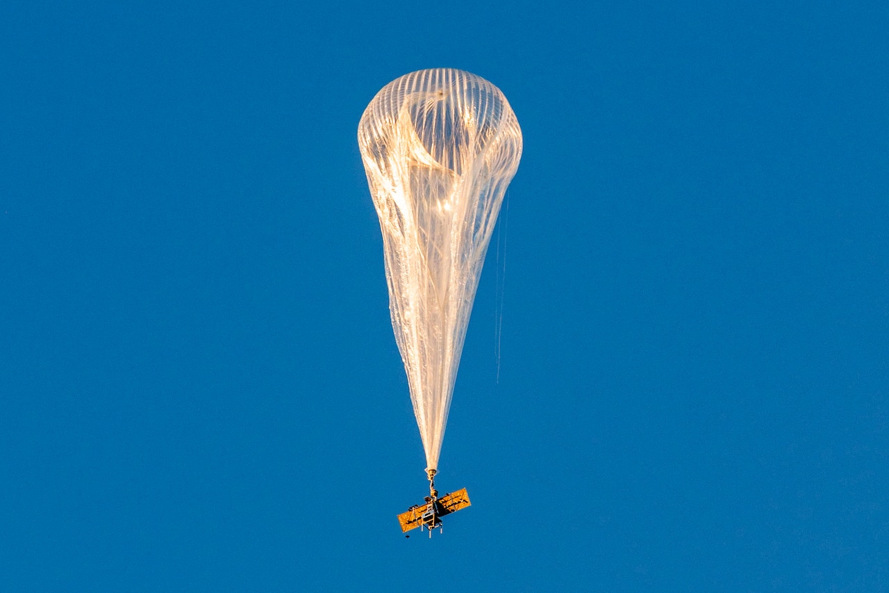 A high-altitude balloon rises against a solid blue sky.