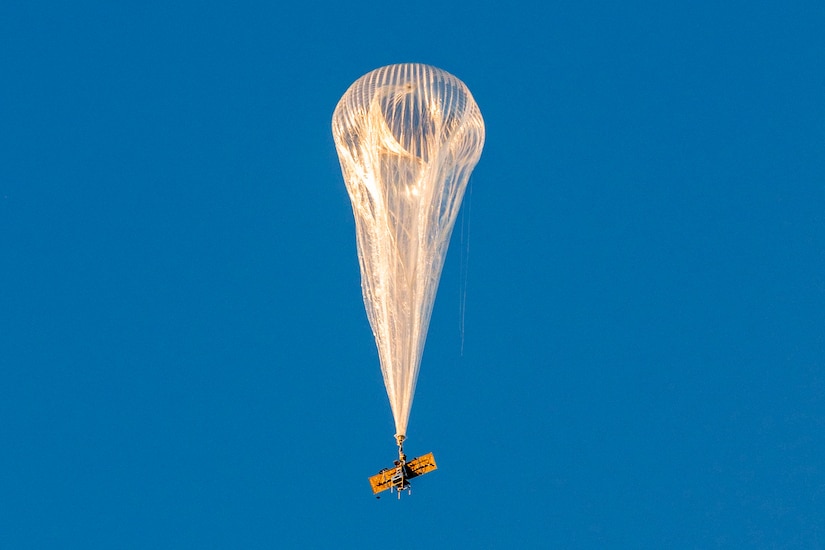A high-altitude balloon rises against a solid blue sky.