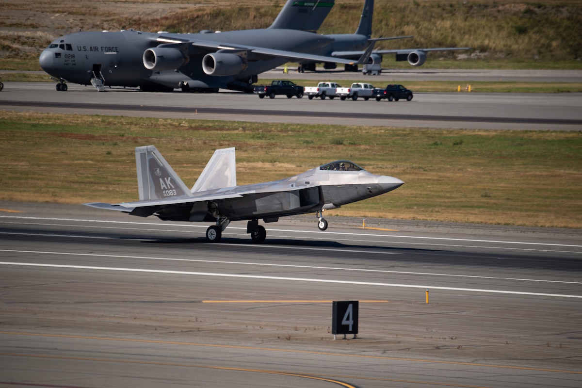 A fighter jet lands on a runway, there is a military transport plane, and four trucks are parked next to it in the background.