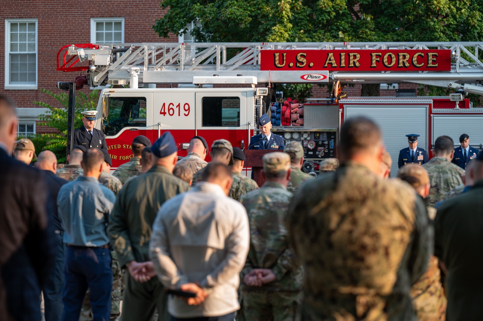 A man in a military dress uniform speaks into a microphone while standing at a podium. Several people in camouflage military uniforms and civilian casual attire bow their heads in the crowd. A red fire truck is parked in the background.