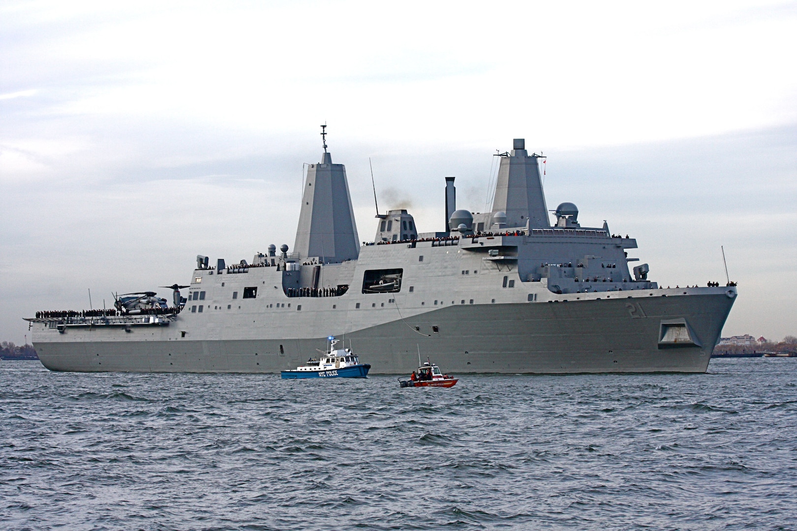 A large gray military ship sails on a river beside a tugboat and a patrol vessel.