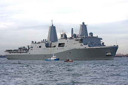 A large gray military ship sails on a river beside a tugboat and a patrol vessel.