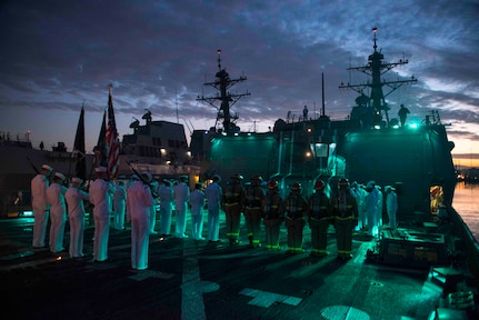Sailors in military dress uniforms and firemen aboard a ship at night bow their heads. The sky is cloudy and there is another large gray ship in the background.