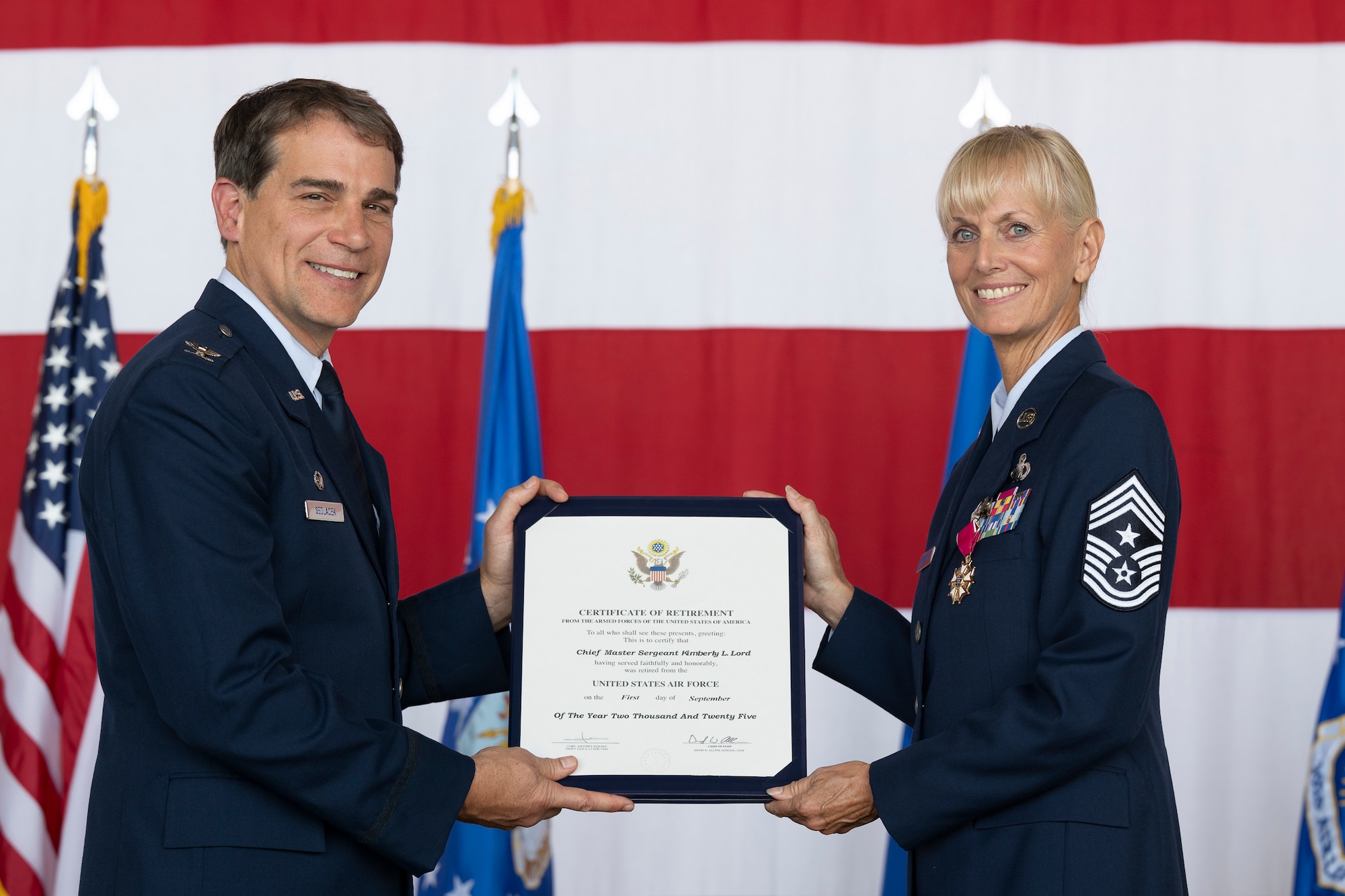 Two military servicemembers hold a piece of paper that reads "CERTIFICATE OF RETIREMENT" with an American flag in the background.