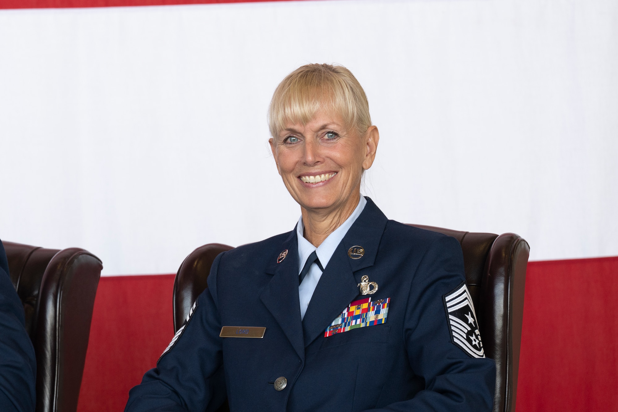 A woman in military service dress smiles while seated with an American flag in the background.
