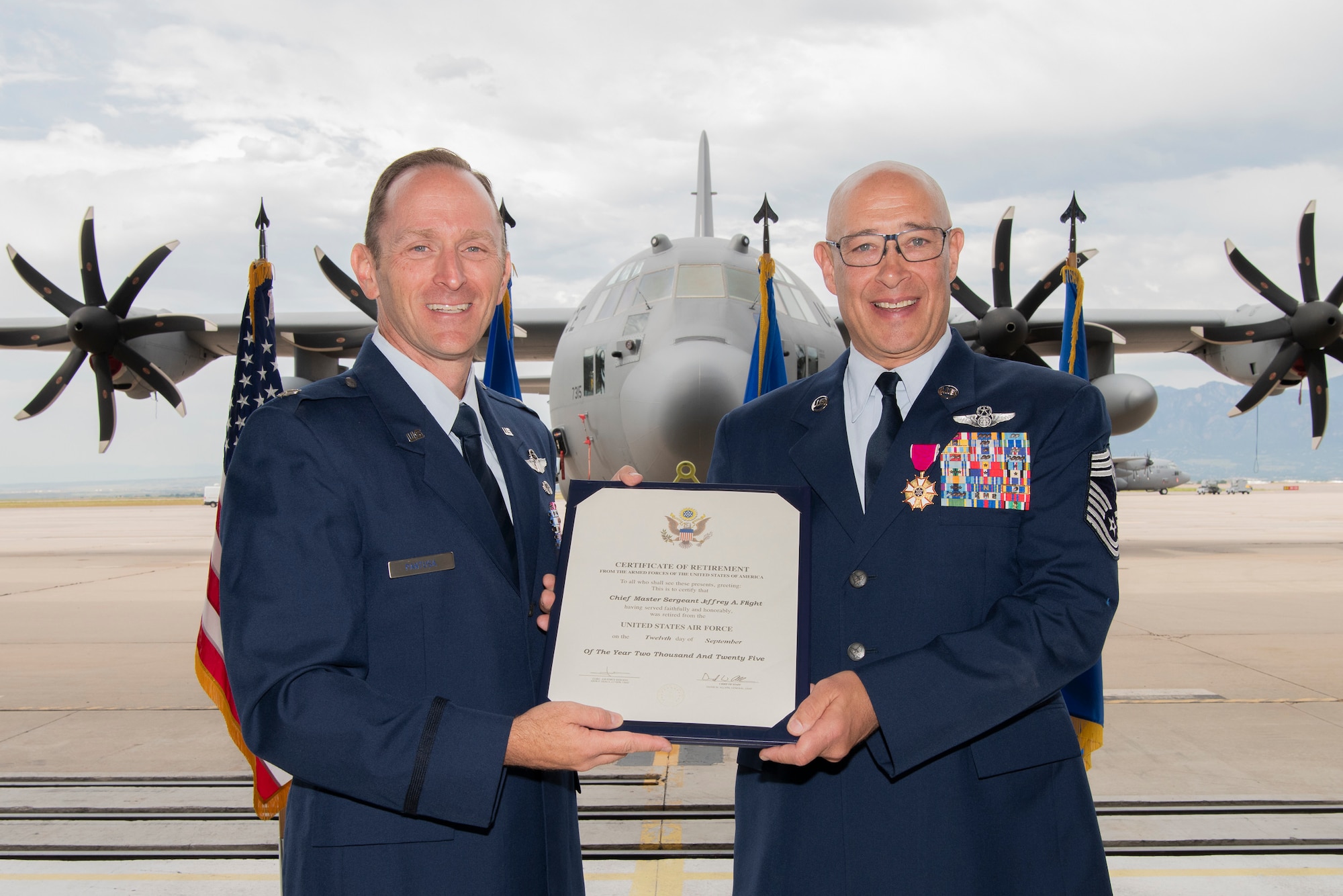 Two men in military service dress hold a piece of paper between them reading "CERTIFICATE OF RETIREMENT" with a military aircraft in the background.