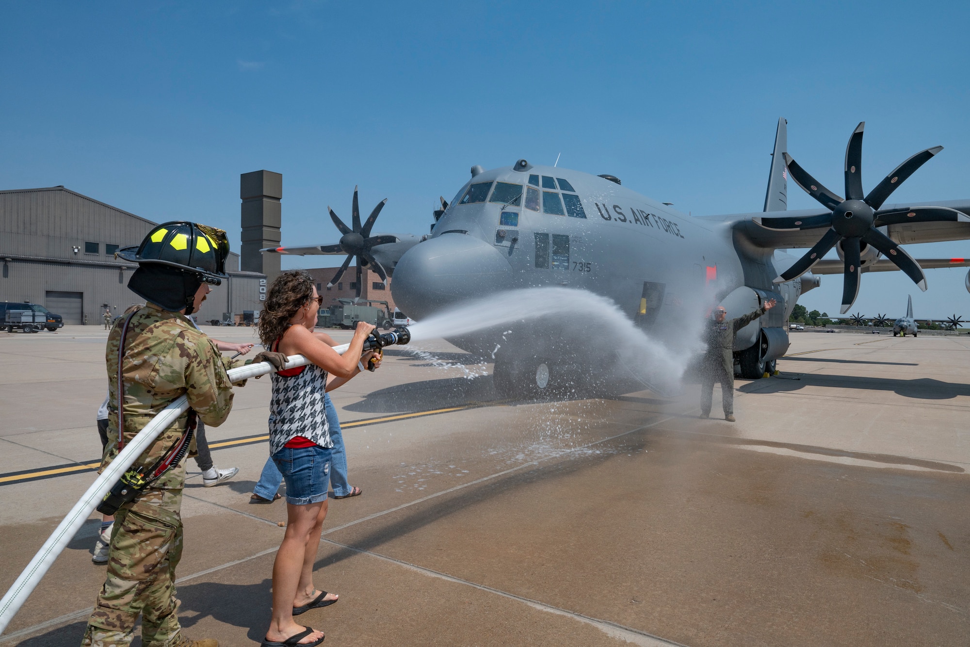 A civilian woman and military firefighter blast a man in a flight suit with water from a hose next to a military aircraft.